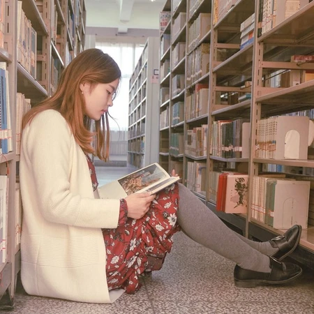 A girl is sitting on the library floor between stalls of books, reading one