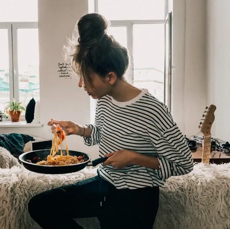 A woman sitting on a couch and eating spaghetti out of a frying pan