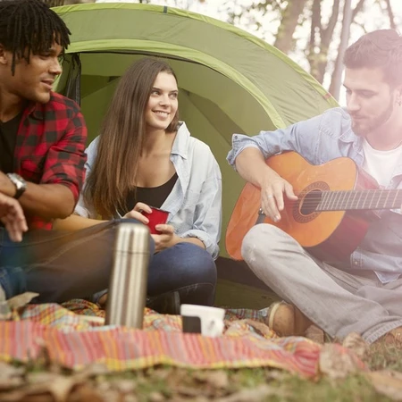 A group of young three young people sitting in the woods next to a tent, one is playing the guitar