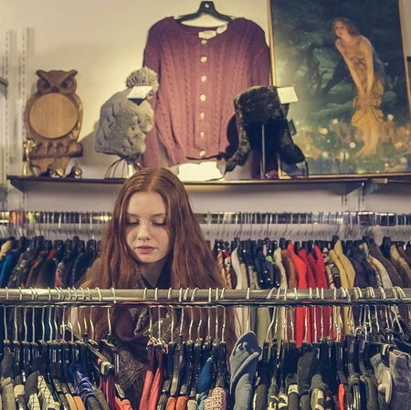 A teenage girl standing between racks of clothing in a second-hand store, hunting for a bargain